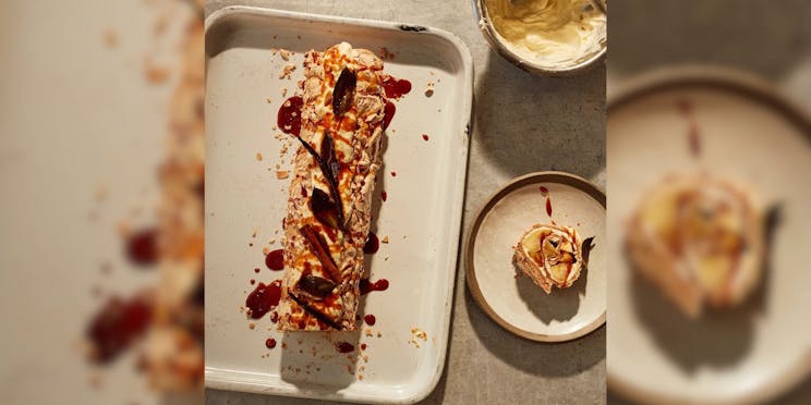 Photo of a meringue roulade on a baking tray, drizzled with burnt honey and a bowl of whipped cream next to it.