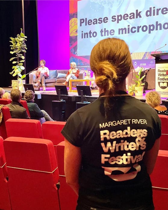 A photo of a volunteer at Margaret River Readers & Writers Festival in the foreground, with a stage with authors talking on it in the background. 