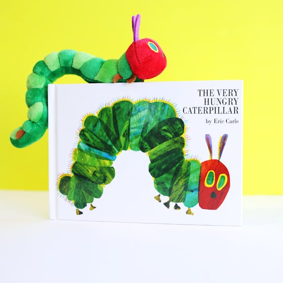 The Very Hungry Caterpillar against a white and yellow background, with a caterpillar toy crawling on the book. 